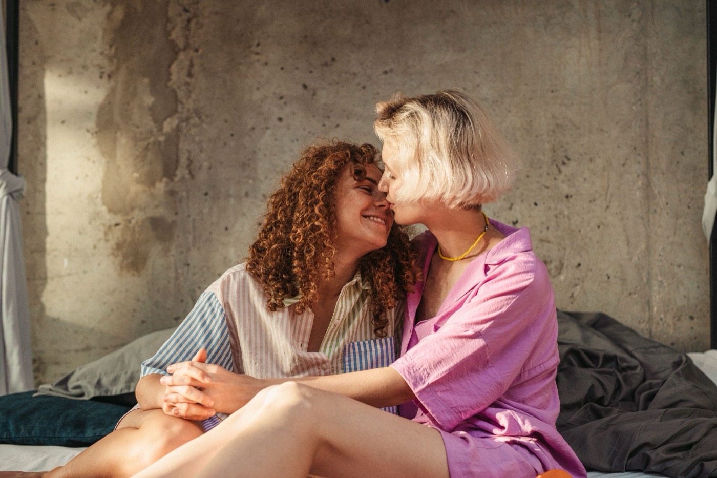 Two women sitting close on a bed in warm sunlight, smiling at each other