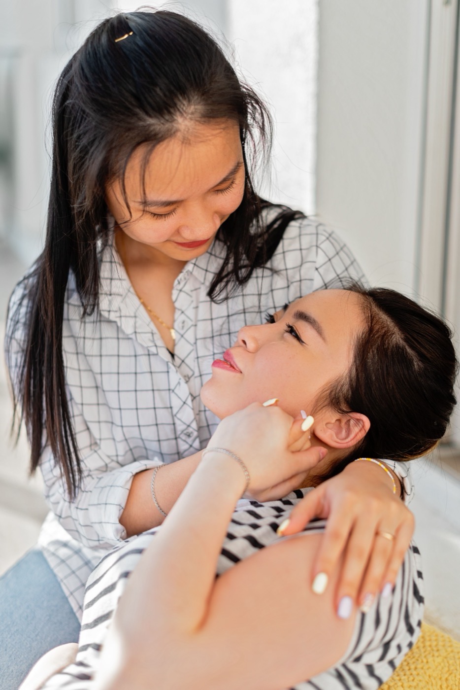 Two women sharing a quiet, affectionate moment beside a bed in soft daylight