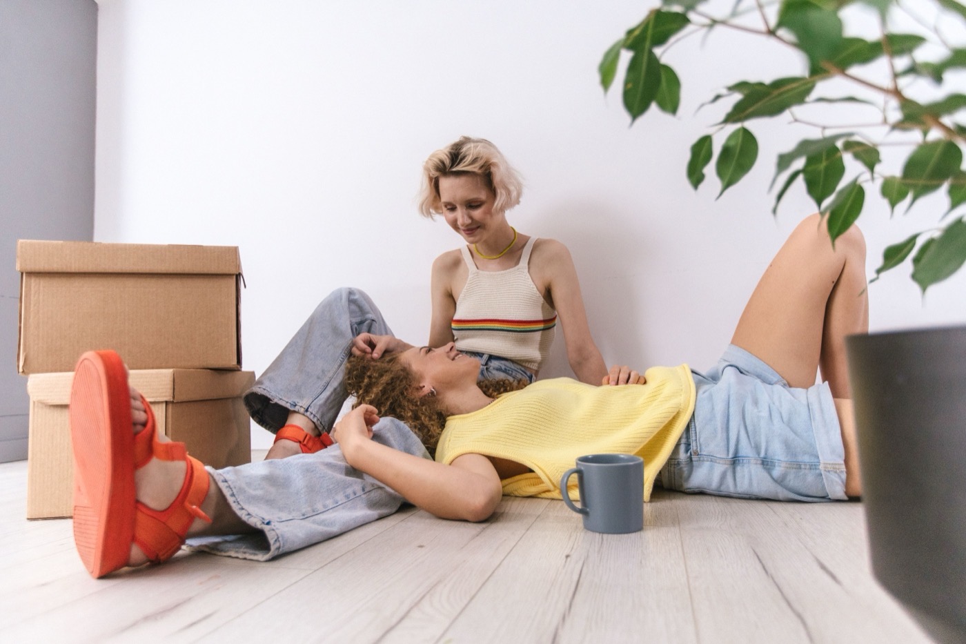 Two women relaxing on a wood floor beside moving boxes, smiling at each other